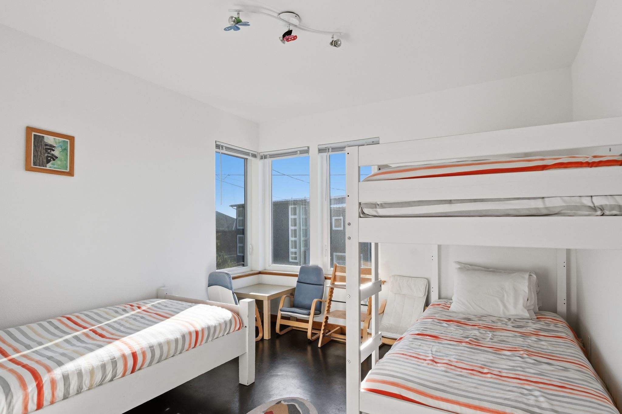 A children's bedroom with white bunk beds and a single twin bed, all with striped bedding. A small table and chairs are by the corner windows, and the room has dark floors and white walls.