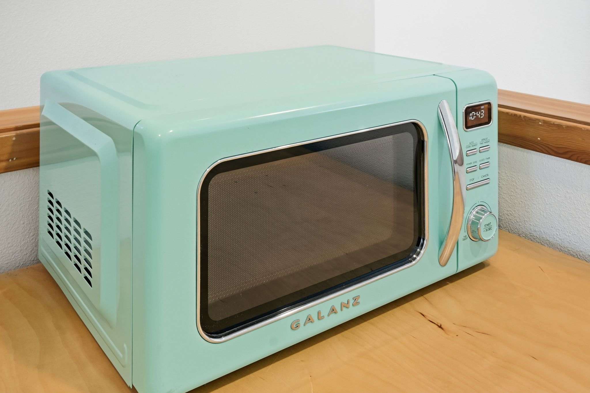 A close-up of a retro-style mint green Galanz microwave with a digital clock displaying "10:43" and a wooden handle, sitting on a light wood countertop.