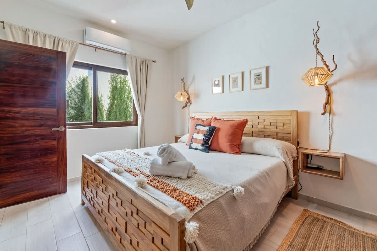 A bright bedroom features a wooden bed frame with a textured white and orange bed runner, flanked by unique branch-like wall sconces. A large wooden door and a window with green foliage outside are visible.
