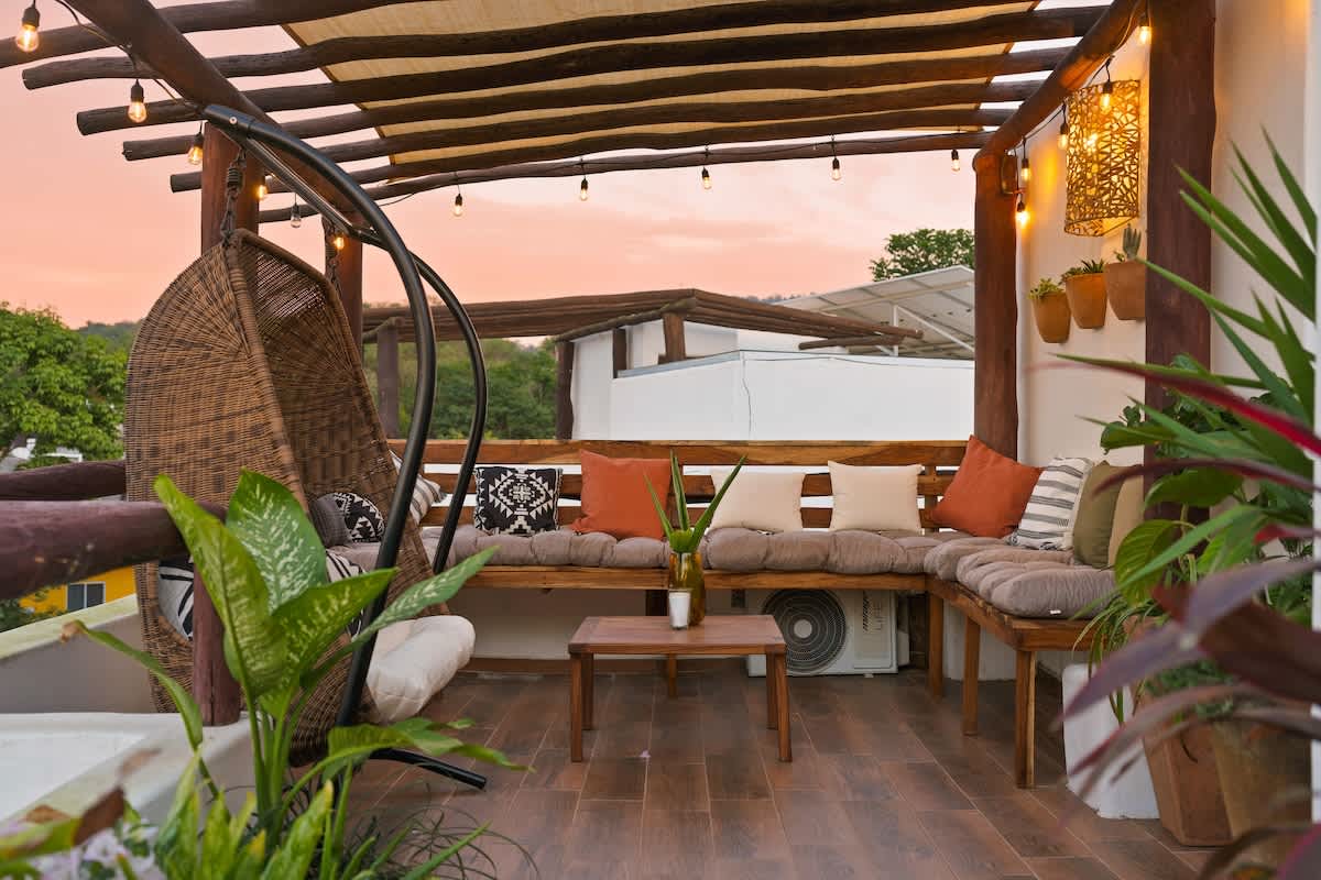 A cozy rooftop terrace features a wooden pergola with string lights, a wicker hanging chair, and built-in wooden benches with cushions, surrounded by potted plants under a warm sunset sky.