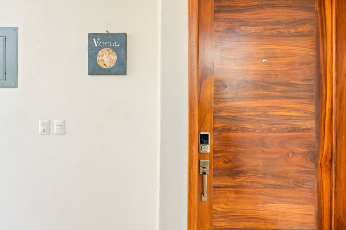 A close-up of a modern wooden front door with a smart lock and handle is shown next to a white wall featuring a small decorative sign labeled "Venus" and electrical outlets. The rich wood grain of the door is prominent.