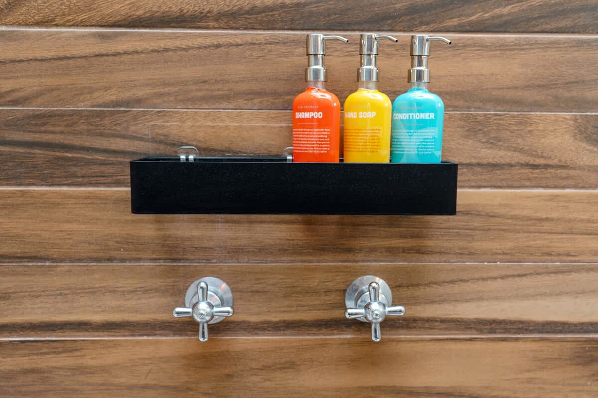 A close-up of a shower wall features three colorful dispenser bottles for shampoo, hand soap, and conditioner on a sleek black shelf, set against a warm, horizontal wood-grain tiled background with chrome fixtures.