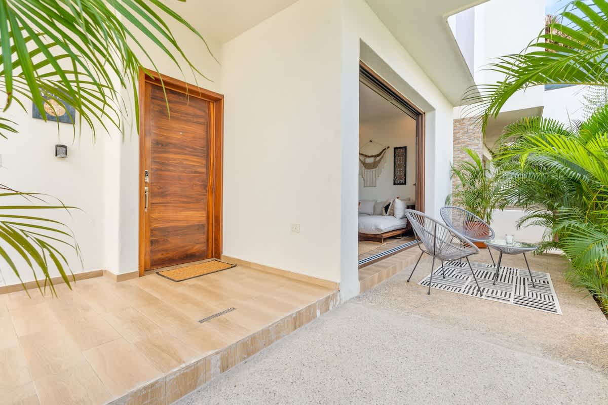 A welcoming entrance features a solid wood door next to an open sliding glass door revealing a bedroom with a bed and decorative wall hanging. A small outdoor seating area with two grey woven chairs and a table sits on the patio, framed by lush palm foliage.