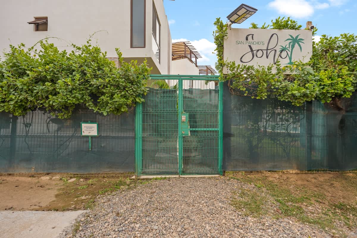 A green metal gate with a lock provides access to the property, flanked by lush green bushes and a white sign reading "San Pancho's Soho" under a blue sky.