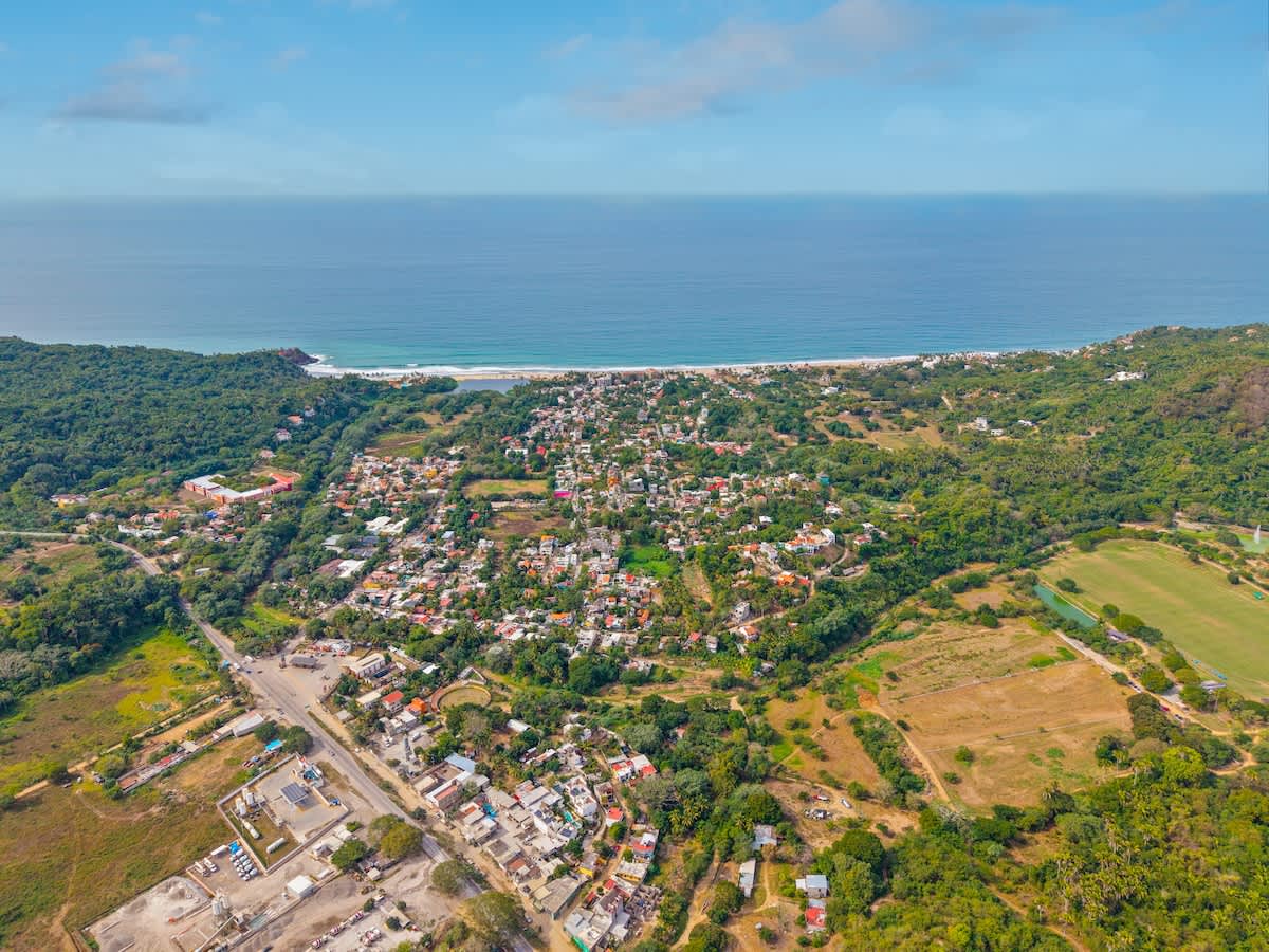 A sweeping aerial view captures a vibrant coastal town nestled between lush green hills and the expansive blue ocean, with numerous buildings scattered among the verdant landscape under a clear sky.