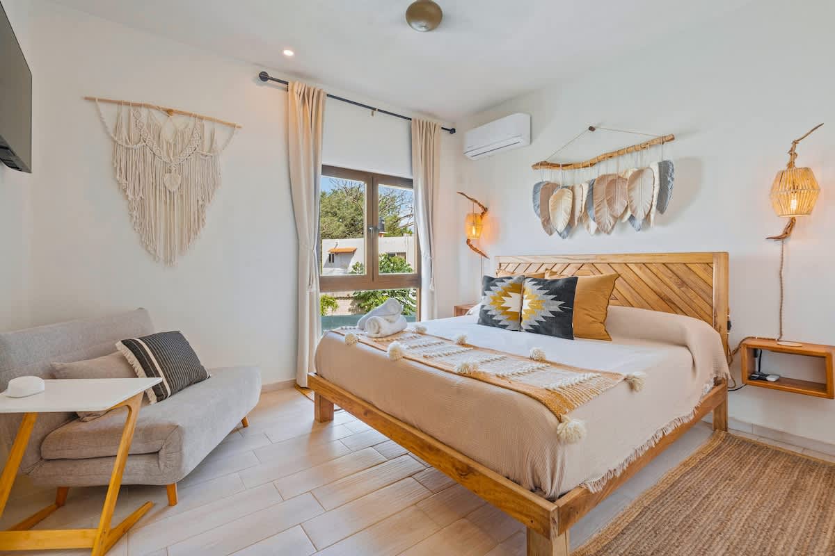 A well-lit bedroom showcases a wooden bed frame with a slatted headboard, adorned with a textured beige throw and a geometric patterned pillow. A small grey sofa with a striped cushion and a side table are positioned near a large window, with a macrame wall hanging above.