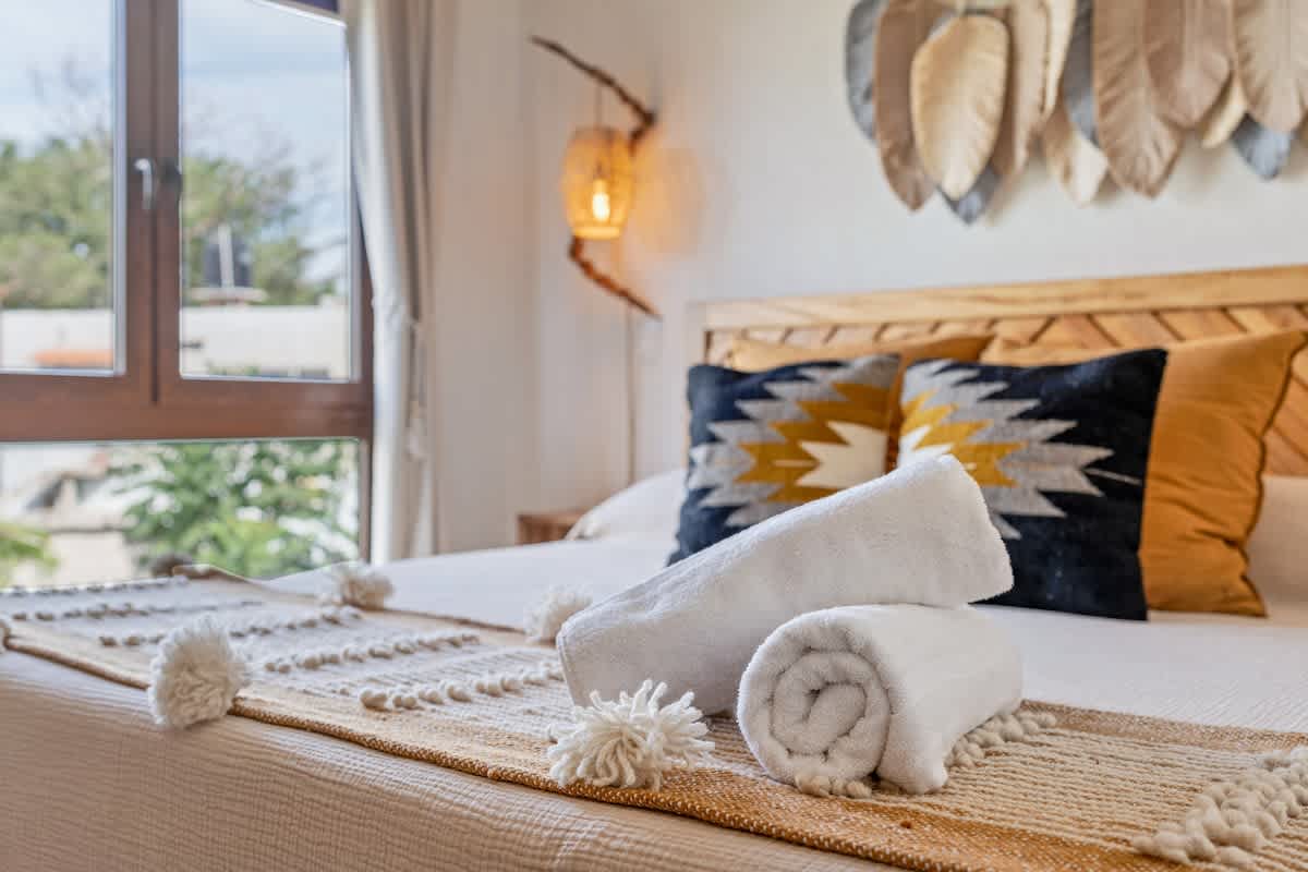 A close-up of a neatly made bed with a white duvet, a textured white and orange bed runner, and two rolled white towels. Decorative pillows with geometric patterns add a pop of color, with a window in the background.