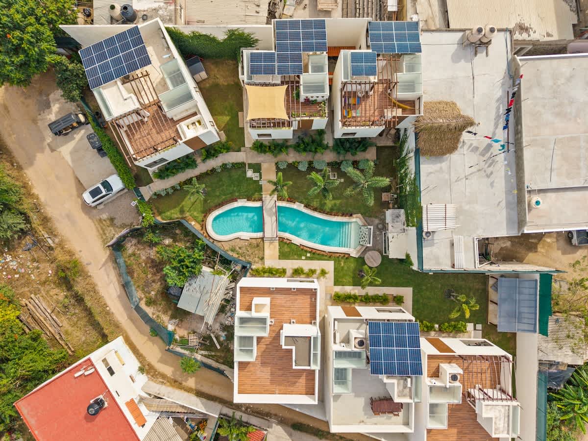 An aerial view showcases a cluster of modern white buildings with solar panels on rooftops, surrounding a winding blue swimming pool connected by a small bridge, all set amidst lush green landscaping.
