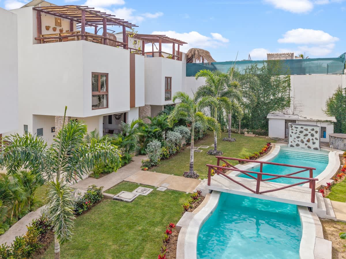The property's exterior showcases modern white buildings overlooking a winding swimming pool with a charming wooden bridge, surrounded by manicured green lawns and tall palm trees.
