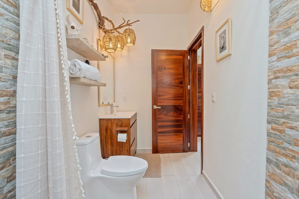 A bright bathroom with stacked stone-look tile in the shower area and a wooden vanity with a white sink. A toilet is visible, and a large mirror reflects the bedroom beyond the open door.