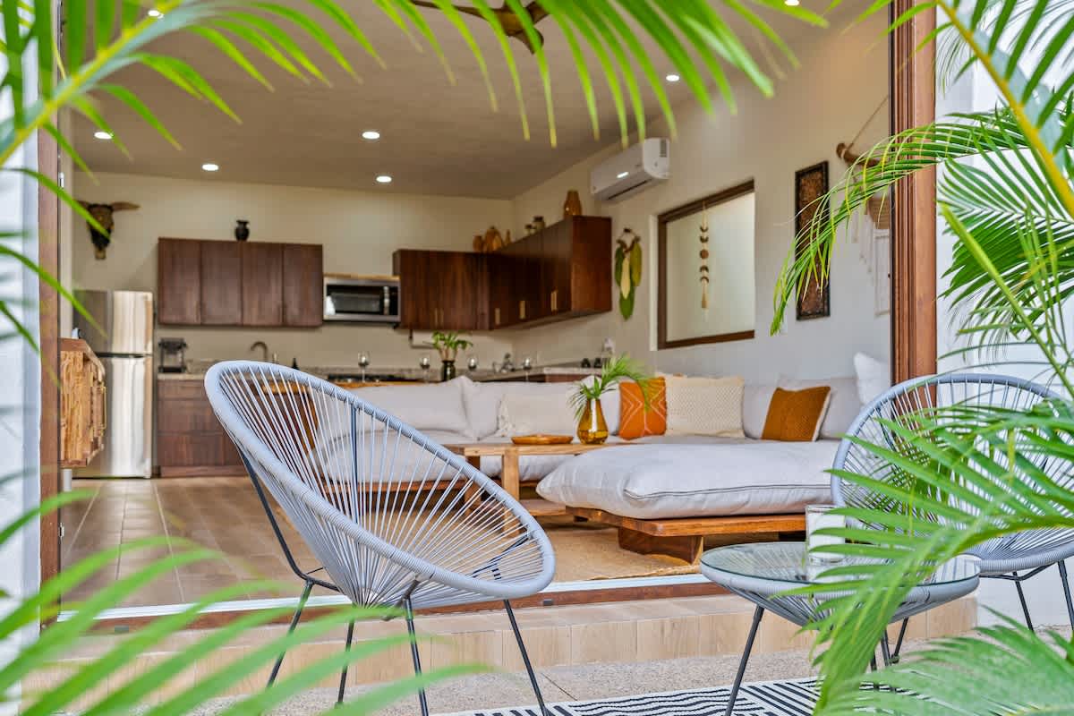 An open-concept living area and kitchen are visible through large sliding glass doors, featuring a light-colored sectional sofa, wooden coffee table, and a kitchen with dark wood cabinetry and stainless steel appliances. Two grey woven outdoor chairs and a small glass table are on the patio in the foreground, framed by lush green palm fronds.