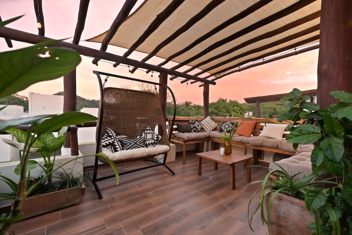 Inviting rooftop terrace at sunset, featuring a large wooden pergola with a fabric canopy, a woven hanging chair, and a long L-shaped bench with cushions, surrounded by potted greenery.