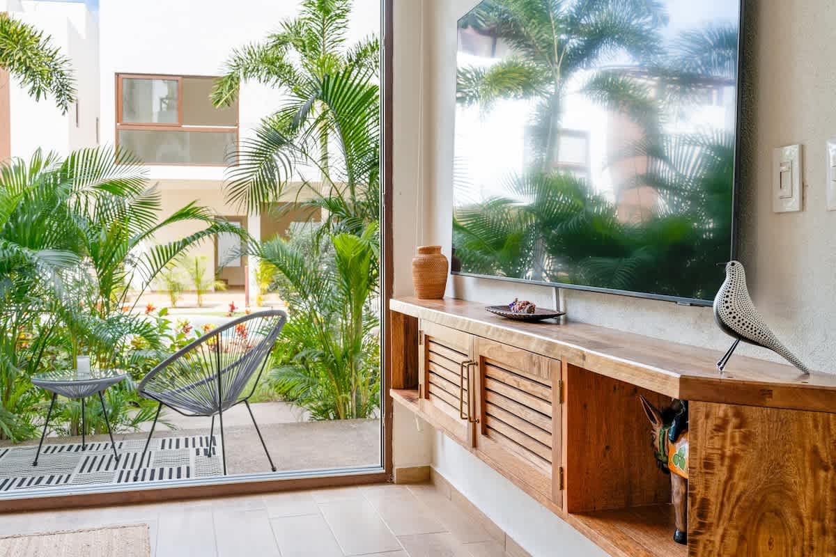 A cozy living space features a large flat-screen TV mounted above a slatted wooden console, with a glass sliding door offering views of an outdoor patio and lush greenery, and a stylish black wire chair.