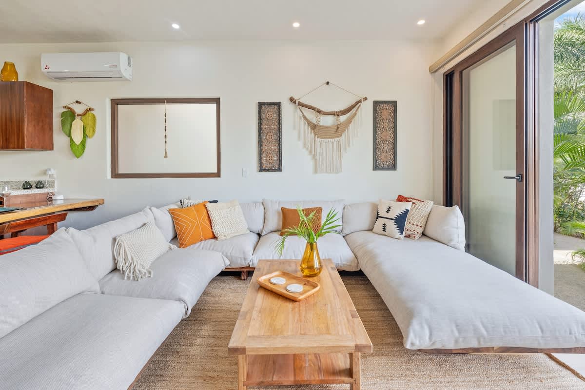 Bright living room featuring a large L-shaped sectional sofa with light upholstery, a wooden coffee table on a jute rug, and a sliding glass door leading to an outdoor patio with lush greenery.