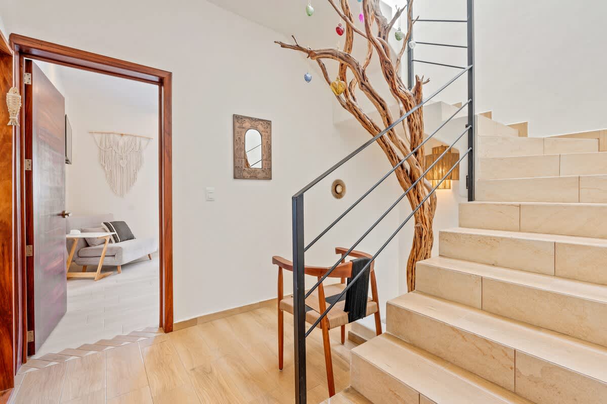 A bright foyer showcases a unique, large decorative tree branch extending upwards alongside a modern staircase with black metal railings and light-colored tiled steps. An open wooden doorway leads to a living area with a macrame wall hanging.