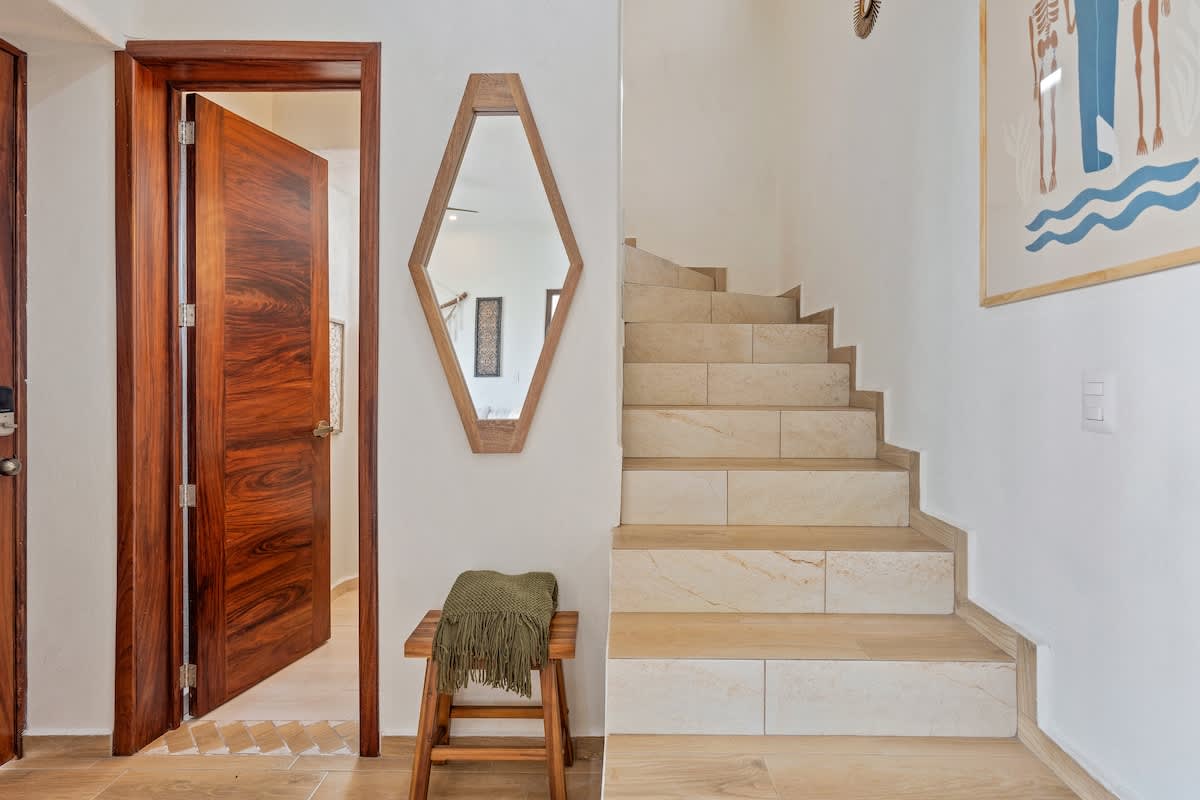 Entryway featuring a wooden door, a unique hexagonal mirror above a small wooden bench with a throw, and a light-colored tiled staircase leading upstairs, with a colorful art piece on the adjacent wall.