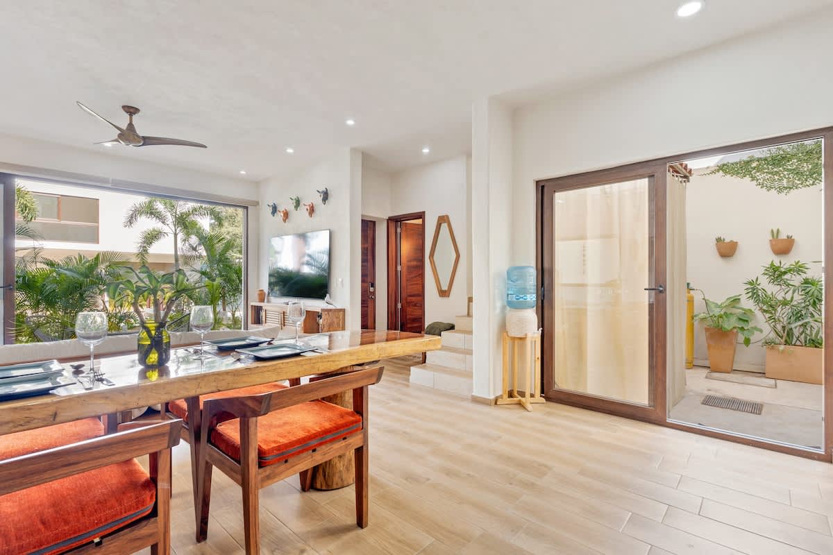 Bright dining area with a large wooden table and orange upholstered chairs, adjacent to a living room with a TV and large windows. A glass door opens to an outdoor patio with plants.