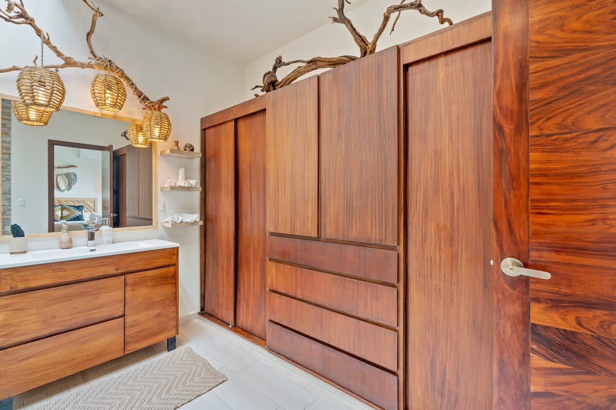 This bathroom features a wooden vanity with a white countertop and sink, illuminated by unique woven pendant lights hanging from decorative branches. A large mirror is positioned above the vanity, and a full wall of rich wooden built-in wardrobes from the adjacent bedroom is visible.