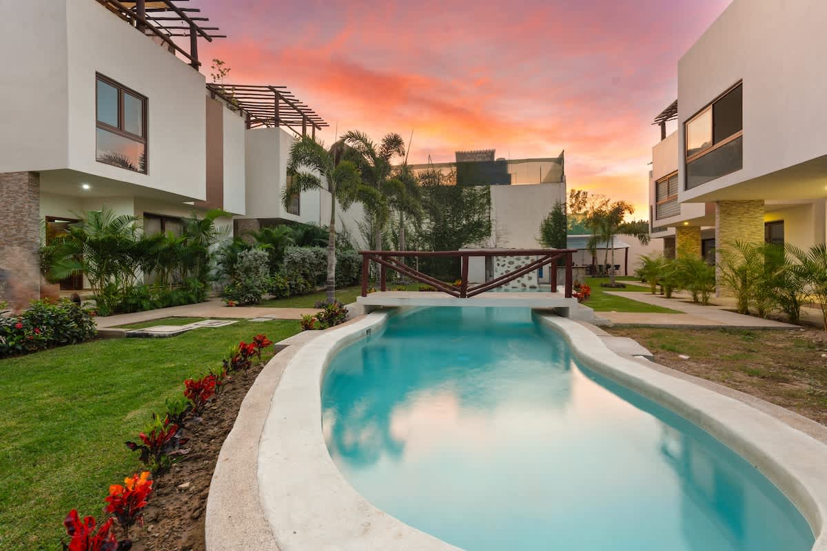 The property's exterior features modern white buildings with a narrow, winding pool in the foreground, connected by a small bridge, all bathed in the warm glow of a sunset.