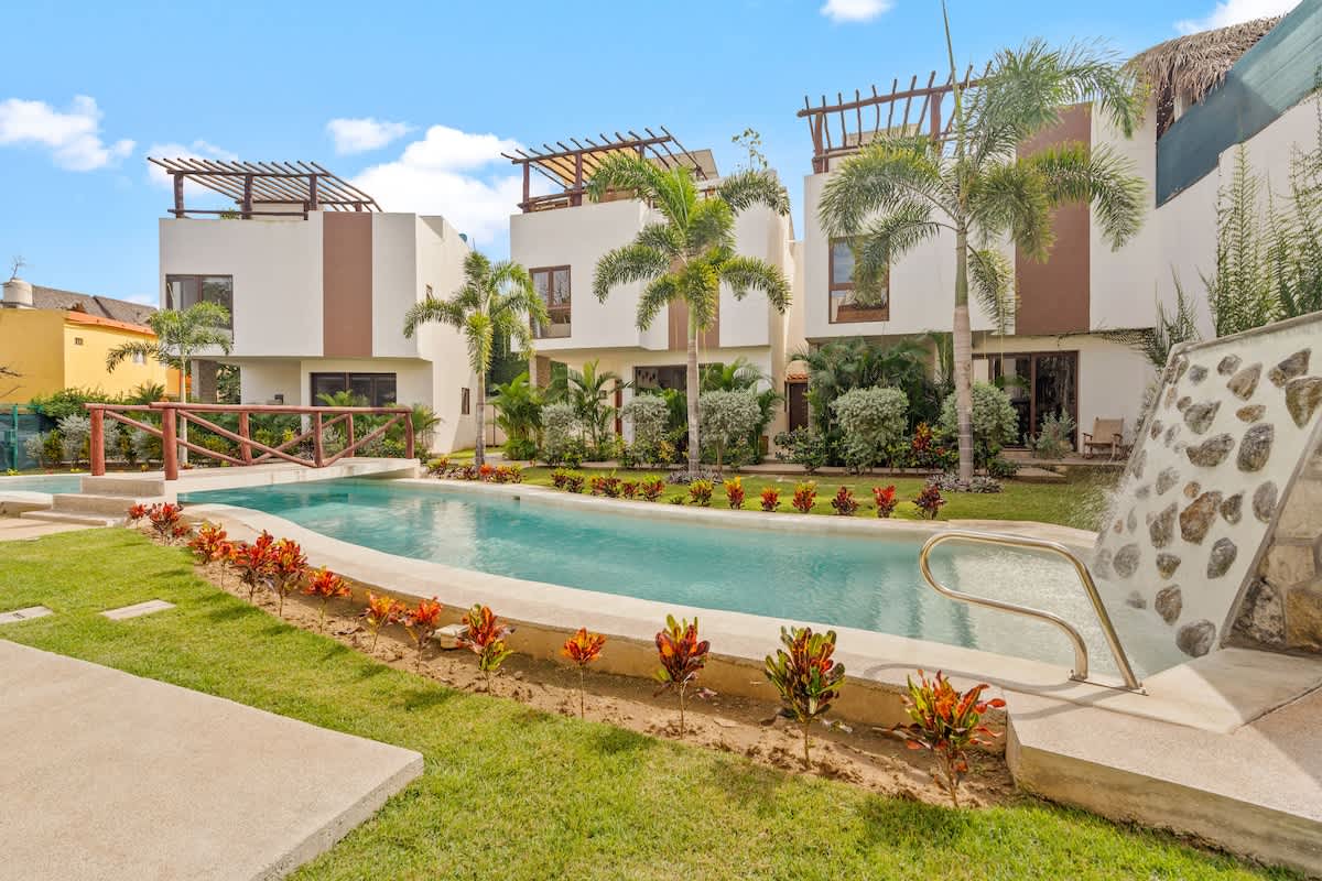 A long, narrow swimming pool with a stone waterfall feature is set within a lush green lawn, bordered by colorful tropical plants, leading to modern white and brown buildings with balconies and palm trees under a bright blue sky. A wooden bridge crosses the pool.