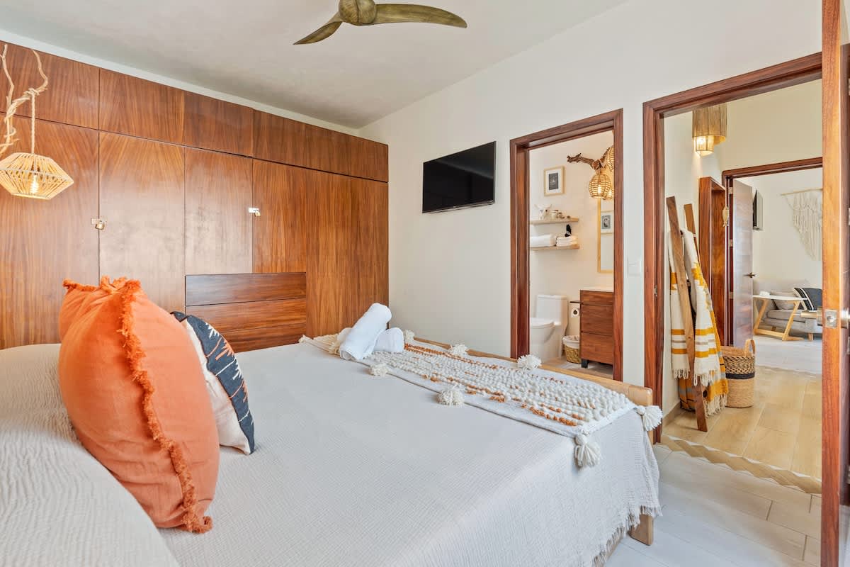 A cozy bedroom with a wooden bed frame and a large built-in wooden wardrobe spanning the wall. Two dark wooden doors lead to an adjacent bathroom and another room, with a wall-mounted TV above the bed.