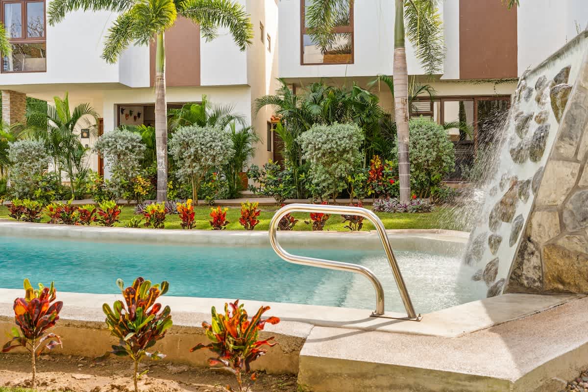 A close-up view of the swimming pool shows clear blue water and a stone waterfall feature with water cascading into the pool, alongside a stainless steel handrail for entry. Vibrant red and green tropical plants line the pool's edge.
