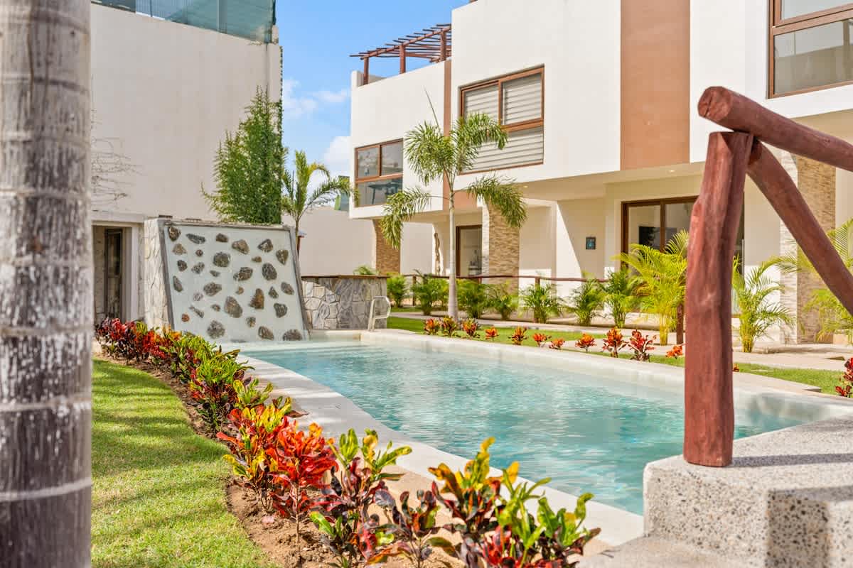 A long, narrow swimming pool with a stone waterfall feature is surrounded by vibrant green grass and colorful tropical plants, leading towards modern white and brown multi-story buildings under a clear blue sky. A wooden railing and concrete steps are visible in the foreground.