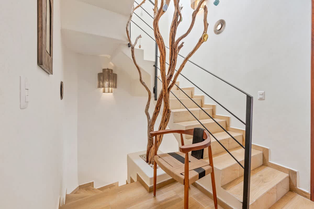 Unique staircase with light-colored steps and a black metal railing, featuring a large decorative tree trunk integrated into the design. A modern chair sits at the base of the stairs.