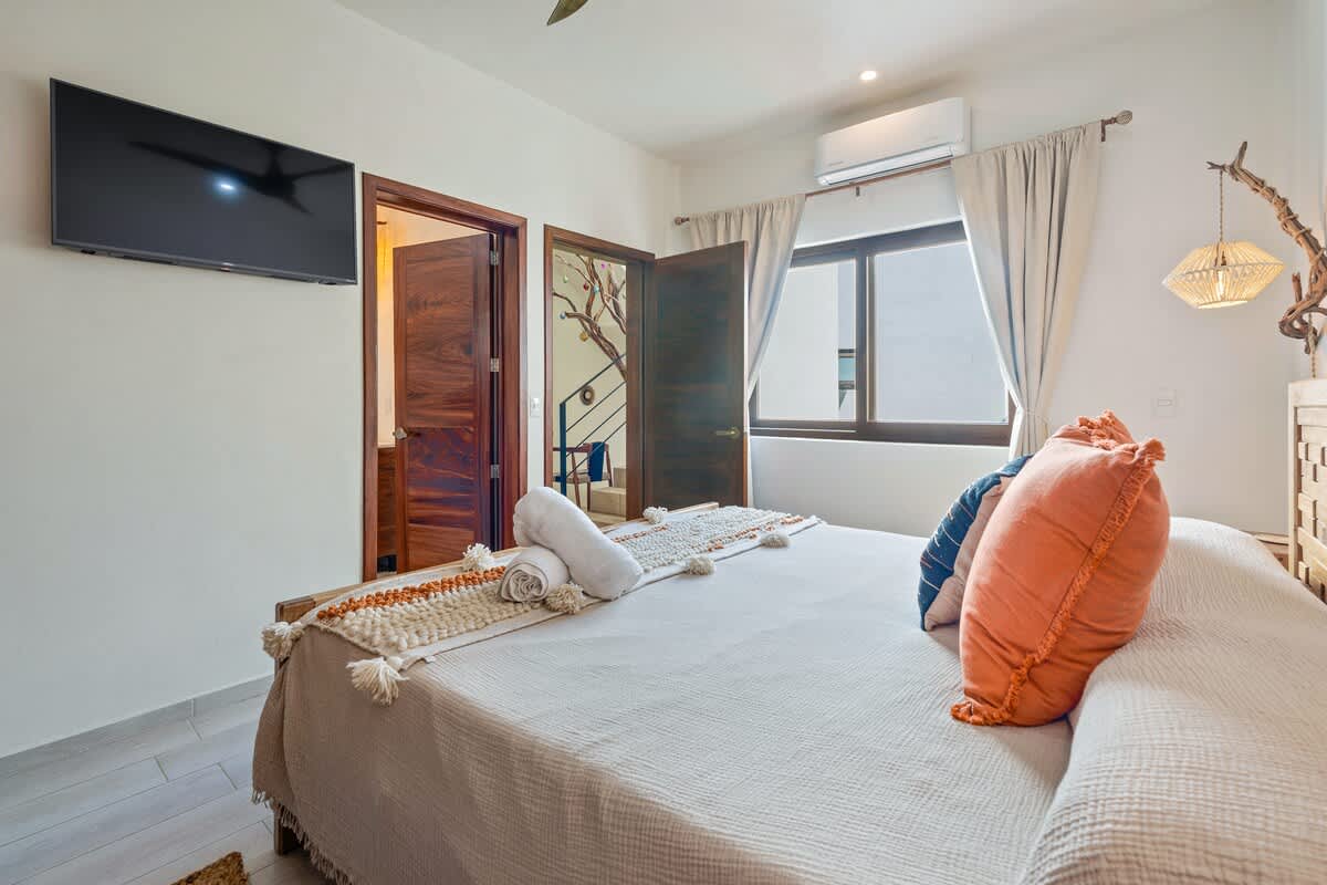 View of a bright bedroom with a wooden bed, a wall-mounted TV, and two dark wooden doors leading to other rooms. The bed is adorned with orange and patterned pillows, and a window offers a glimpse of the outdoors.