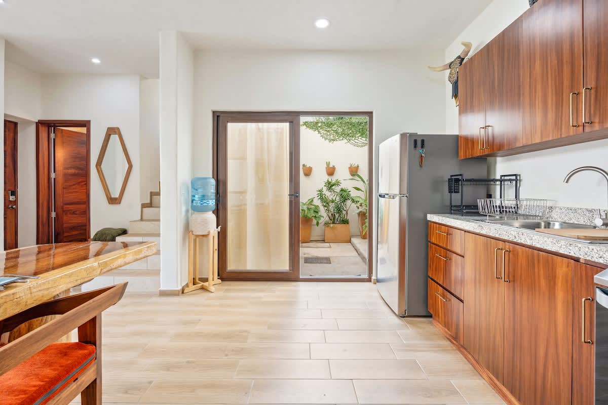 Open-concept kitchen with wood cabinets and granite counters, leading to a dining area with a wooden table and a view of a small outdoor patio through a glass door. A stainless steel refrigerator is visible.
