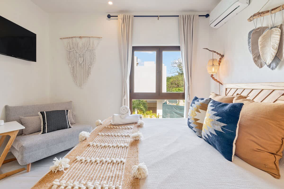 This bright bedroom includes a wooden bed frame with a patterned accent pillow and a textured throw, facing a wall-mounted television. A small grey sofa with a striped pillow sits beside the bed, and a large window provides natural light.