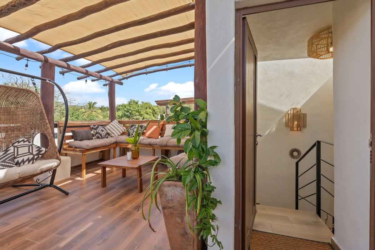 View from the top of an interior staircase leading to a bright rooftop deck, featuring a wooden pergola with a fabric canopy, outdoor seating, and a hanging chair, with lush greenery visible outside.
