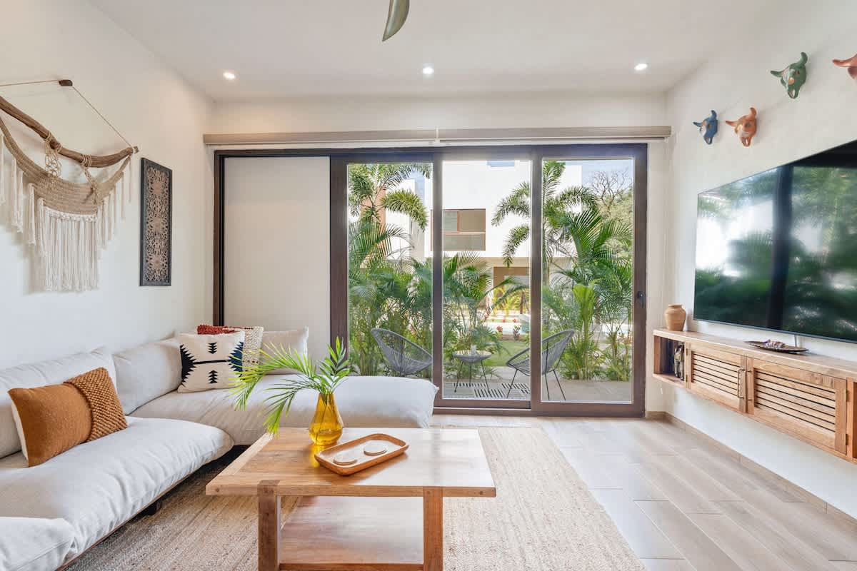 Cozy living room with a light-colored sectional sofa, a wooden coffee table, and expansive sliding glass doors offering a view of a lush tropical garden and an outdoor seating area.