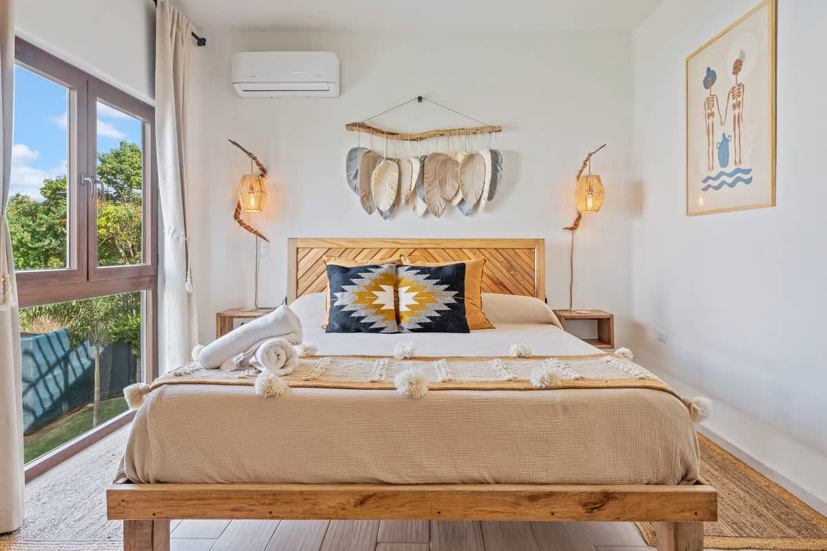 A cozy bedroom features a light wood bed frame with a slatted headboard, dressed with a beige and white pom-pom throw and a patterned accent pillow. Large windows offer views of lush greenery, while a unique feather-like wall hanging and woven pendant lights adorn the wall.