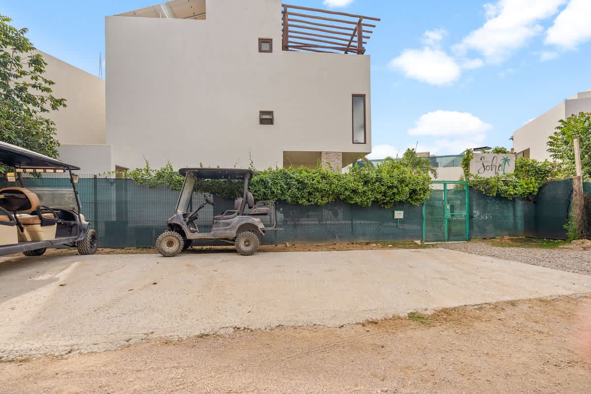 A dark golf cart is parked on a gravel lot next to a green mesh fence covered in vines, with a modern white building visible in the background under a partly cloudy sky.