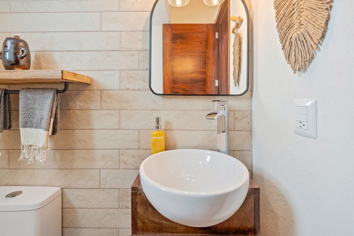 A modern half-bathroom features a white vessel sink on a dark wood vanity, complemented by a sleek chrome faucet and a rounded rectangular mirror. The walls are clad in light-colored rectangular tiles, with a white toilet partially visible on the left.
