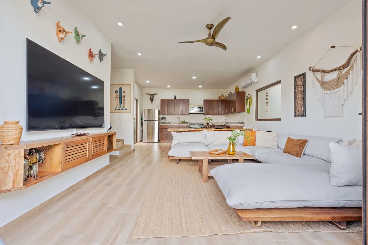 Spacious open-concept living area with a large flat-screen TV mounted above a wooden console, a comfortable L-shaped sectional sofa, and a view into the modern kitchen with dark wood cabinetry.
