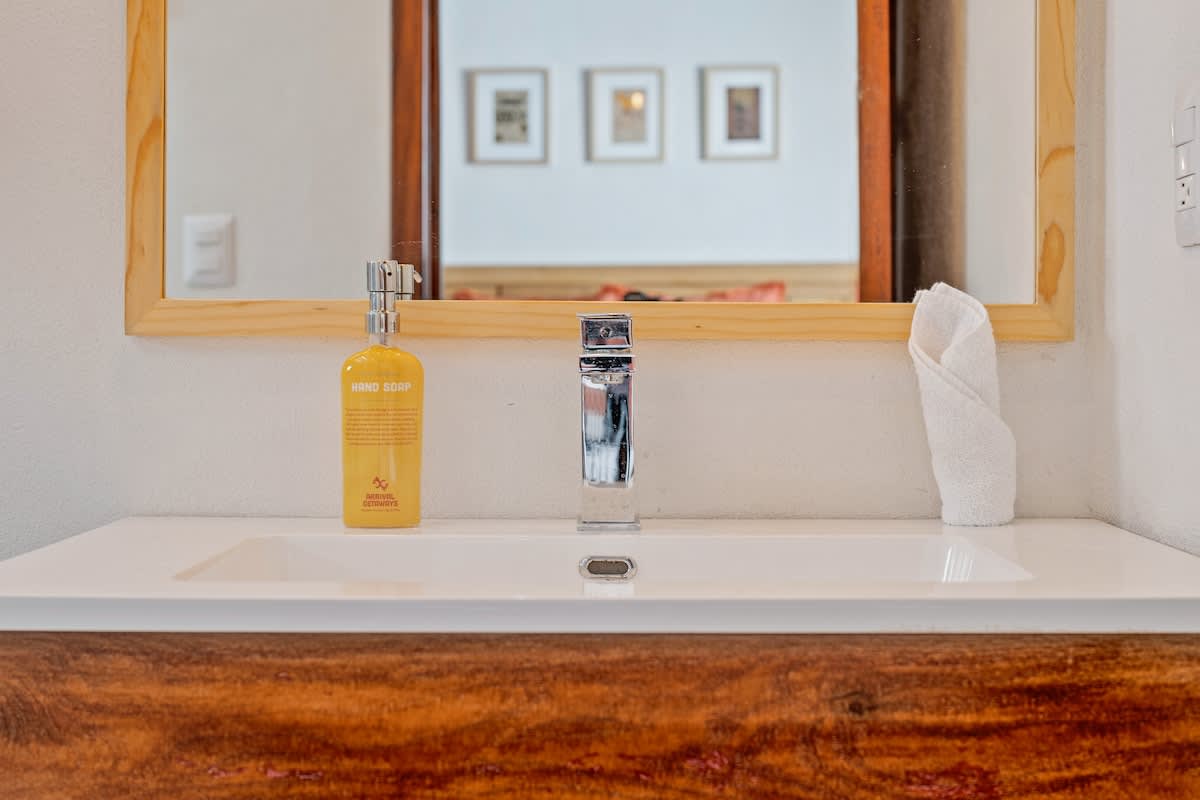 A clean bathroom vanity features a white rectangular sink with a contemporary chrome faucet, a light wooden-framed mirror, and a yellow hand soap dispenser, with a neatly folded white towel on the counter.