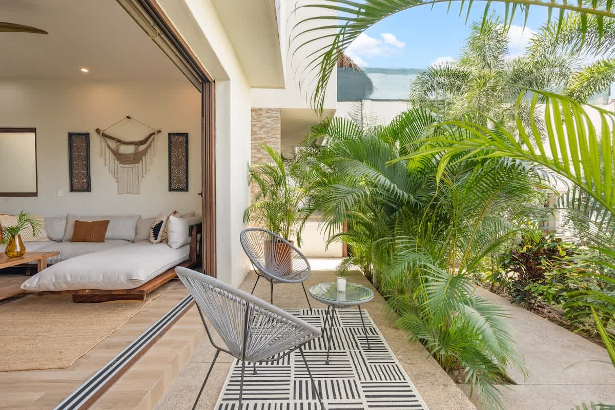 Private outdoor patio featuring two modern grey string chairs and a small round table on a geometric black and white rug, surrounded by lush tropical plants and accessible from the living room.