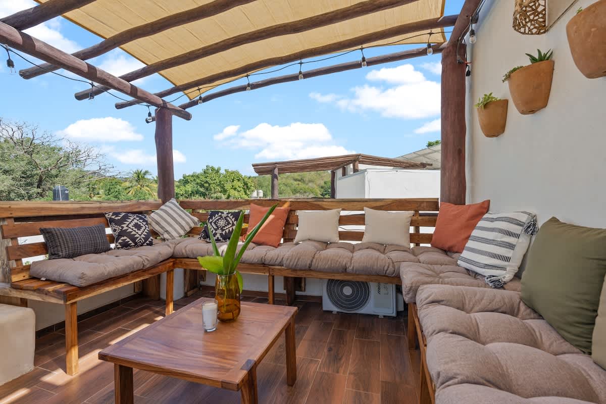 A spacious rooftop deck with a large L-shaped wooden bench seating area adorned with cushions and pillows. A wooden coffee table sits in the center, all under a shaded pergola with string lights.