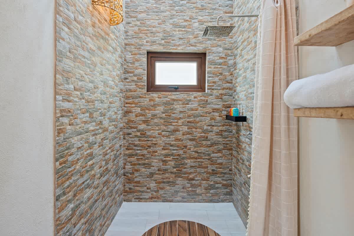 A modern shower stall with stacked stone-look tile walls and a small horizontal window. A wooden slatted mat covers part of the floor, and a shower curtain is visible to the right.