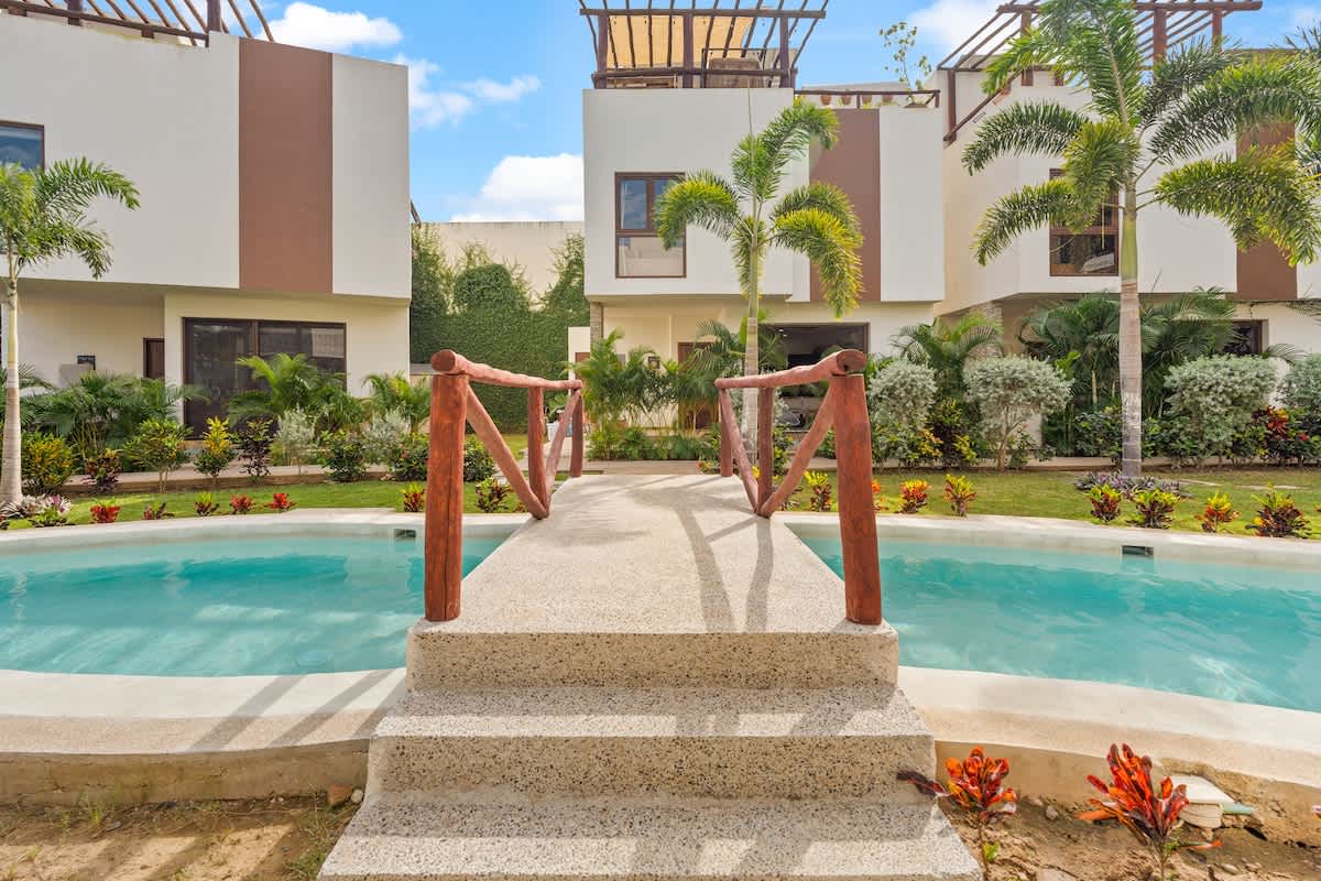Modern white buildings with brown accents flank a narrow, winding pool, connected by a rustic wooden bridge. Lush green landscaping and palm trees surround the area under a bright sky.