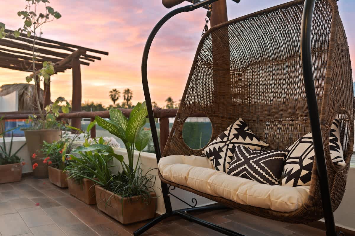 A close-up of a comfortable wicker hanging chair with patterned pillows on a rooftop terrace, overlooking a vibrant sunset sky with distant palm trees.