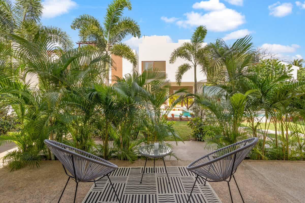 Secluded outdoor patio area with two contemporary grey woven chairs and a small glass-top table on a striped rug, enveloped by dense, lush tropical foliage and palm trees.
