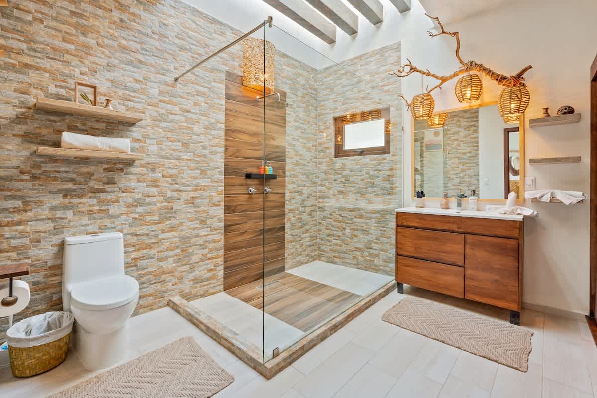 A spacious bathroom features a large walk-in shower with glass enclosure and wood-look tile accent wall, alongside a white toilet and a double wooden vanity with two vessel sinks. The walls are clad in textured stone-look tiles, and woven pendant lights hang above the vanity.