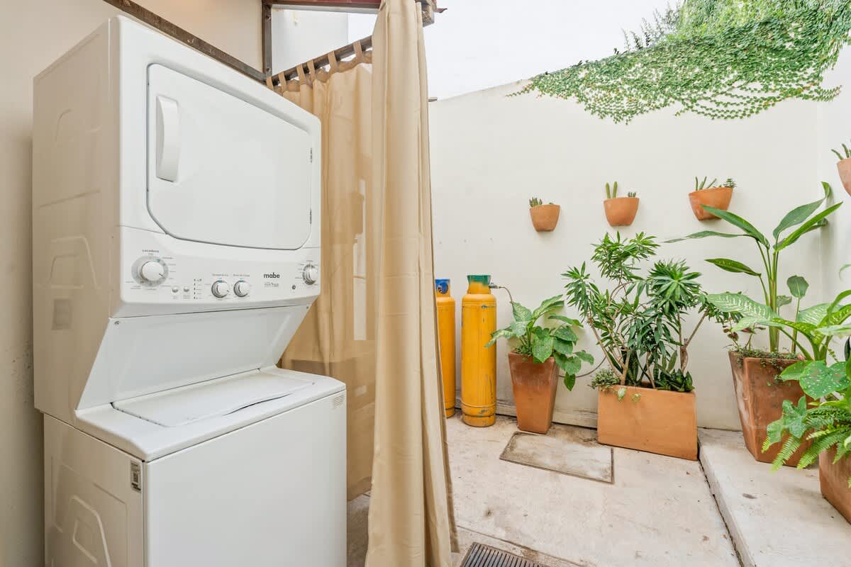 Outdoor laundry area on a concrete patio, featuring a stacked white washer and dryer unit. Potted plants and wall decor are visible in the background.