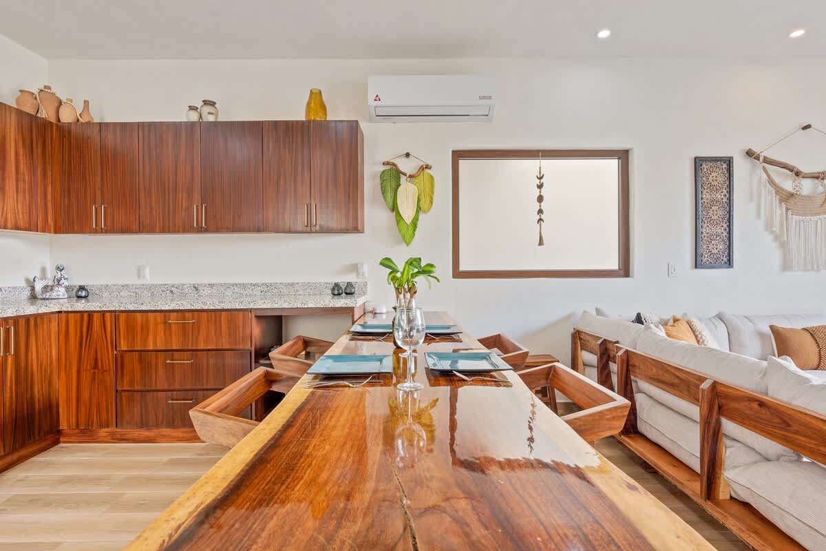 Dining area featuring a striking live-edge wooden table set with place settings and wine glasses, surrounded by wooden chairs with orange cushions, adjacent to the modern kitchen and living room.