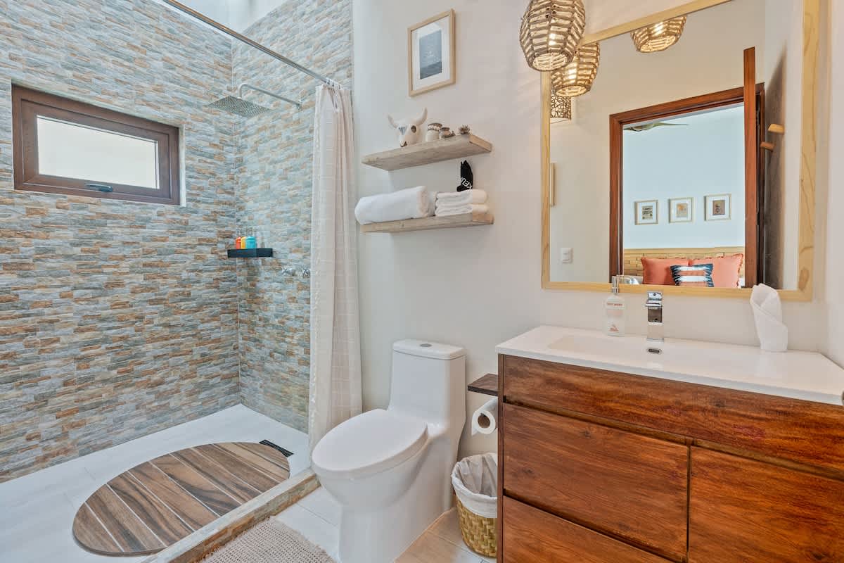 A contemporary bathroom featuring a shower with stacked stone-look tile walls and a wooden vanity with a white sink. A toilet is positioned next to the shower, and a large mirror reflects the bedroom.