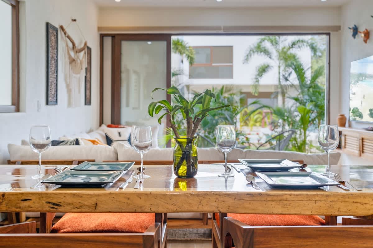 Detailed view of a polished wooden dining table set with square plates and wine glasses, featuring a green vase with foliage, with the living room and outdoor greenery visible beyond.