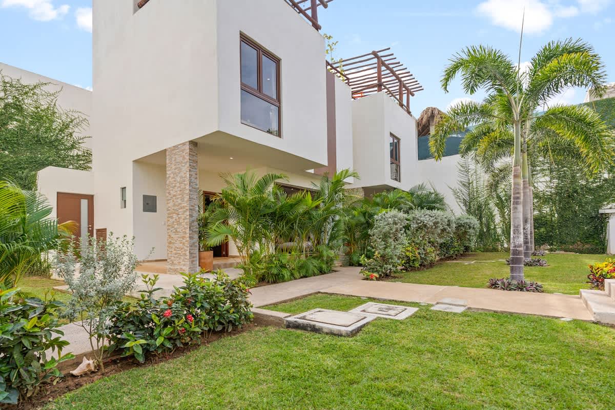 Modern white multi-story buildings with brown accents, set amidst vibrant green lawns and abundant tropical landscaping, including tall palm trees and various shrubs.